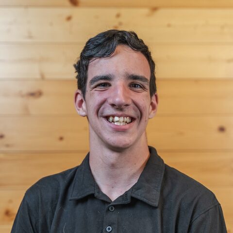 Russ Gohl smiling, in a black polo shirt in front of a light wood-paneled wall